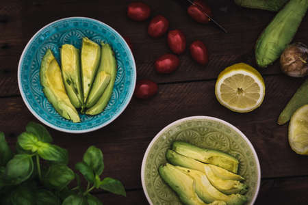 Two bowls with sliced avocado, tomatoes and lemon on a dark wooden table as preparation for an appetizer salad. Top view.の写真素材