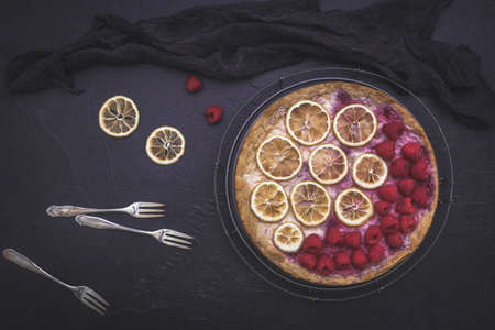 Cheese cake with raspberries, dried lemon slices, forks and black cloth on dark background, top viewの写真素材