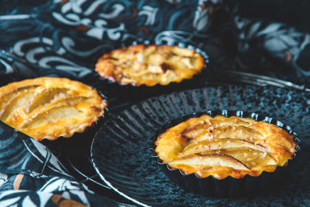 Three baked apple tartlets in black tart dishes on a black plate with black and white patterned...