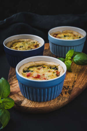 Baked vegetable tartlets in blue colored tart dishes on a wooden board on dark blue background....