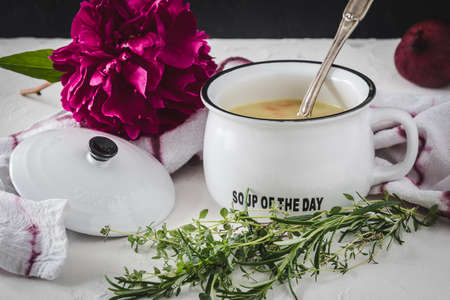 Potato soup in white bowl with handle and lid on white table, decorated with red onions, purple peony blossom and herbs.の写真素材
