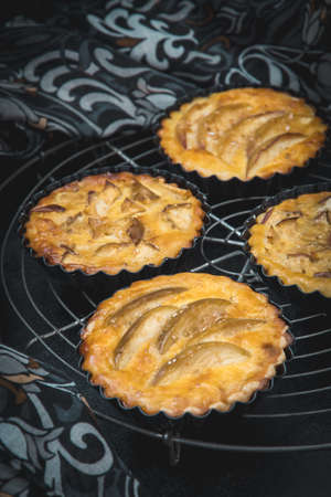 Baked apple tartlets in black tart dishes on a cooling rack with patterned cloth. Vertical closeup.