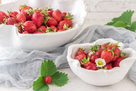 Handmade white pottery bowls with fresh strawberries on white background, decorated with strawberry leaves and gray clothの写真素材
