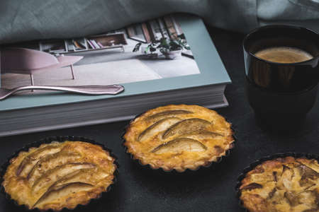 Apple tartlets and a coffee on a black table prepared for a break. In the background a coffee table book and a cloth, moody and pastel colors.の写真素材