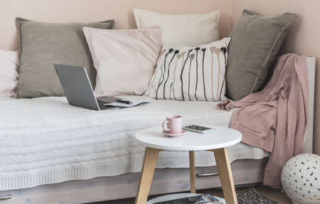 Cozy room with bed, gray and rose pillows, coffee table, notebook, mobile phone and coffee cup prepared for a break, interior in Scandinavian styleの写真素材