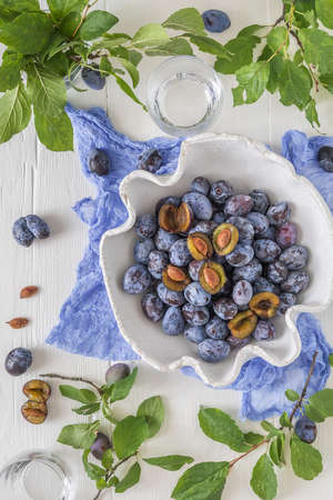 White curved bowl filled with ripe plums, some of them sliced, on a white wooden table with green leaves. Top view.の写真素材