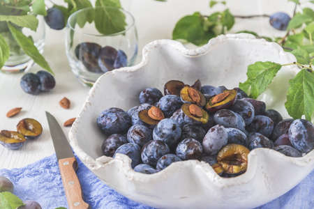 White curved bowl filled with ripe plums on a white wooden table with green leaves, some of them sliced. Horizontal closeup.の写真素材