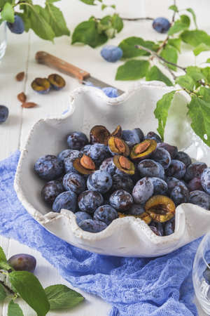 White curved bowl filled with ripe plums, some of them sliced, on a white wooden table with green leaves. Vertical stock photo.の写真素材
