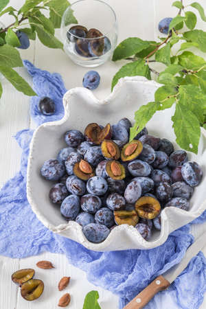 White curved bowl filled with ripe plums, some of them sliced, on a white wooden table with green leaves. Vertical stock photo.の写真素材