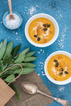 Two white bowls with pumpkin / carot soup served with cream and pumpkin seeds on blue background, decoreated with sage and salt. Vertical top view.の写真素材