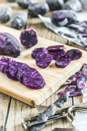 Peeled and sliced purple potatoes on a wooden board, whole potatoes in the background, verticalの写真素材