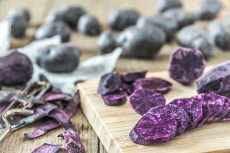 Peeled and sliced purple potatoes on a wooden board, whole potatoes in the backgroundの写真素材