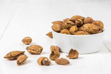 Roasted and salted snack almonds with shell in a small white bowl on white wooden background, copy spaceの写真素材
