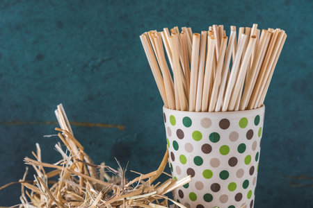 Natural straw drinking straws in a bamboo beaker and a bundle of straw on an old wooden beam, green background. Concept of sustainability, no plastic. Closeup with copy space.の写真素材