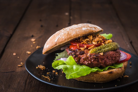 Burger with vegetarian hamburger patty, salad, tomatoes and roasted onions on a black plate on wooden table, copy spaceの写真素材