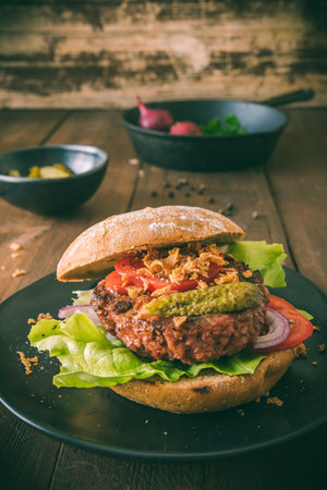 Vegetarian burger with salad, cucumber, tomatoes and roasted onions on a black plate on wooden table, vertical stock photoの写真素材