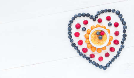 Heart symbol made of different berries with a creme brulee in the middle on white wood, top view with copy spaceの写真素材