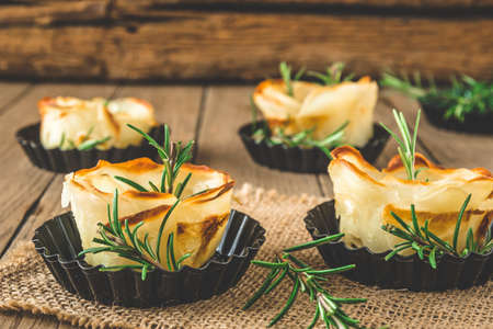 Baked crispy potato roses with rosemary in black tartlet forms on a rustic wooden tableの写真素材