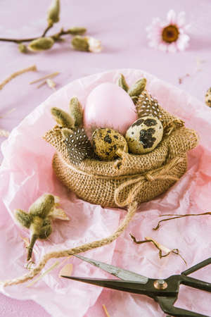 Scene of handmade Easter decoration: jute fabric, pink Easter egg, natural quail eggs, feathers and branches on pinkish background, verticalの写真素材