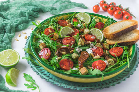 Bowl of fresh vegetable salad: arugula, fresh and dried cherry tomatoes, pine nuts and cheese on a white wooden backgroundの写真素材