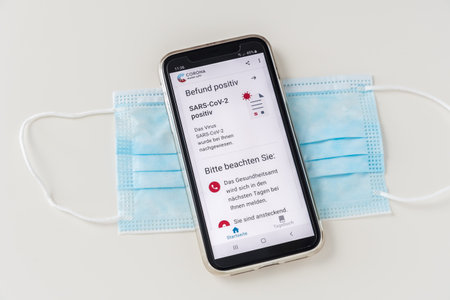 Germany - March 27th 2021: Smartphone with a positive Covid-19 SARS-CoV-2 test result in the official German Corona warning app of Robert Koch institute on a protective mask, white background. Result positive = result positive, Please note = Please note.のeditorial素材