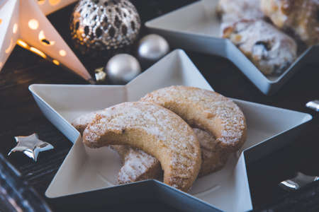 Vanilla-flavored crescent cookies on a white star plate on dark wooden tray, decorated with christmas balls, silver stars and starlightの写真素材