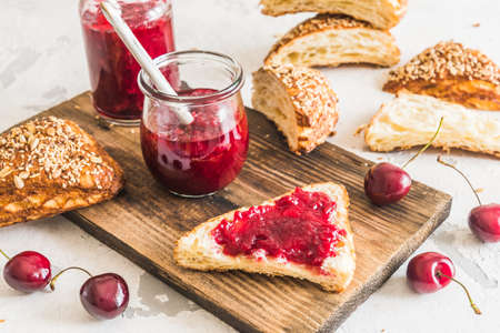 Homemade cherry jam, fresh cherries and cut lye rolls on a wooden board on a white tableの写真素材