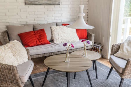Rattan sofa with two armchairs and cozy red and white pillows, white stone wall behind, scandinavian style.の写真素材