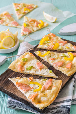 Pieces of tarte flambee with salmon, yellow paprika and spring onions on a wooden board on light green background, verticalの写真素材
