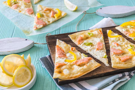 Pieces of tarte flambee with salmon, yellow pepper and spring onions on a wooden board on light green backgroundの写真素材