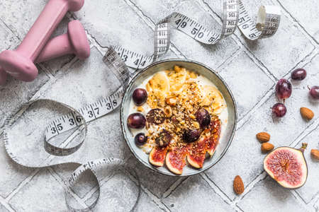 Bowl of yoghurt, fruits and nuts for a healthy diet breakfast or snack. Pink dumbbells and a tape measure on gray background, weight loss concept. Top view.の写真素材