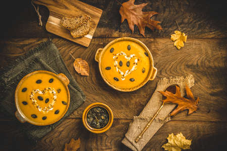 To bowls with pumpkin soup with cream and seeds on a rustic metal and wooden background, decorated with autumn leaves, top viewの写真素材