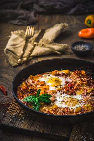 Shakshuka in an iron pan on wooden background. Middle eastern traditional dish. Fried eggs with tomatoes, bell pepper, vegetables and herbs. Vertical.の写真素材