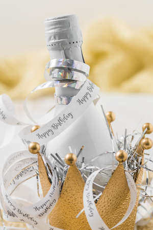 Small white bottle with sparkling wine in a decorative golden metal crown, decorated by streamers with text Happy Birthday. Vertical close up.の写真素材