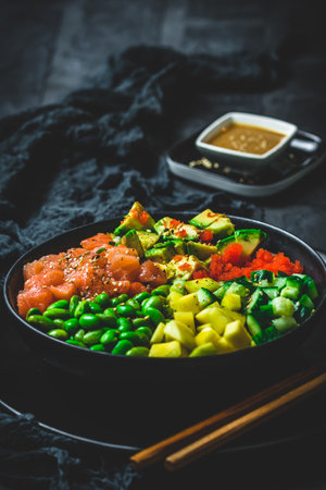 Japanese poke or buddha bowl with salmon, avocado, roe, mango, pies and cucumber in a black bowl on black background, verticalの写真素材