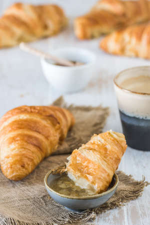 Piece of delicious French croissant dipped in jam on a white wooden table, a cup of coffee and croissants in the background, breakfast scene, verticalの写真素材
