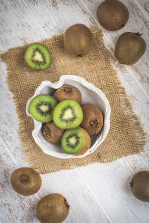 Whole and halfed kiwis in a bowl and on a white wooden table, top viewの写真素材