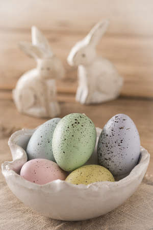 Decorative Easter eggs in pastel colors in a white handmade bowl on a wooden table, two white wooden Easter bunnies in the backgroundの写真素材