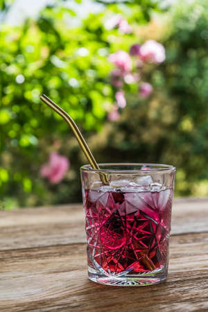 Beautiful ornamental glass with delicious red fruit juice or cocktail with golden metal straw on a wooden garden table, verticalの写真素材