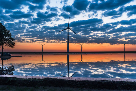 Wind turbines in the warm sunset light with a beautiful reflection in the water and cloudy sky.