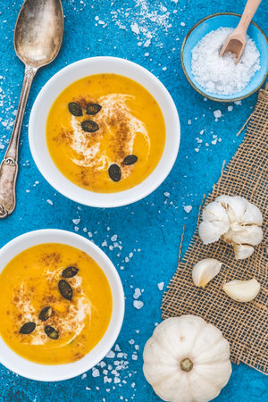 Pumpkin / carrot soup served in two white bowls with cream and pumpkin seeds on a blue background. Vertical top view.の写真素材