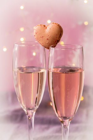Two glasses with sparkling wine with a heart shaped macaron in between, pink pastel background with fairy lights, romantic atmosphere, verticalの写真素材