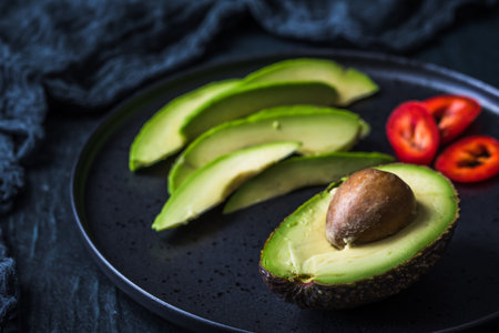 Half avocado with pit and avocado slices on black plateの写真素材