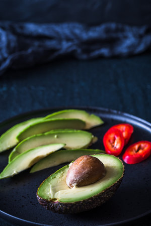 Half avocado with pit and avocado slices on black plate, vertical with copy spaceの写真素材