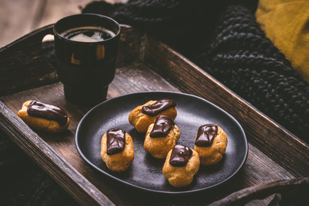 Mini eclairs with dark chocolate and a coffee on a wooden tray, french pastry specialty with custard creamの写真素材