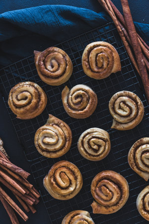 Homemade cinnamon rolls on a cooling rack with cinnamon sticks and dark blue cloth, top viewの写真素材