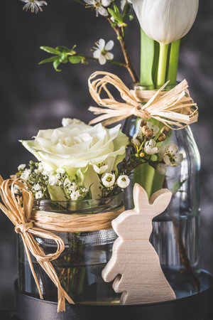 Easter decoration with white rose and tulip and wooden bunny on a black metal stand, gray background, verticalの写真素材