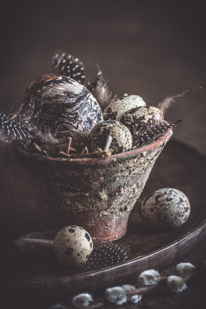 Easter decoration: rustic flower pot with natural quail eggs and feathers on a dark wooden plate and table, vertical with copy spaceの写真素材