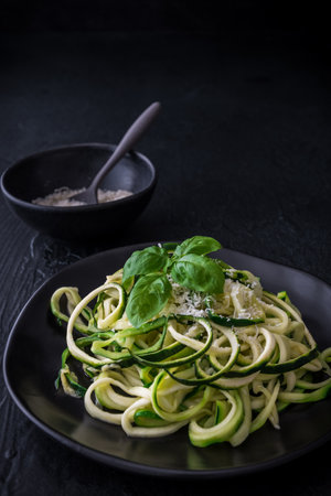Zoodles, zucchini noodles with parmesan and basil on a black plate on black background. For low carb, keto, vegetarian, paleo nutrition and diet. Vertical with copy space.の写真素材