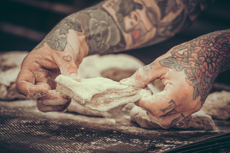 A baker with tattoos kneads bread dough in the bakery. Vintage style with grain.